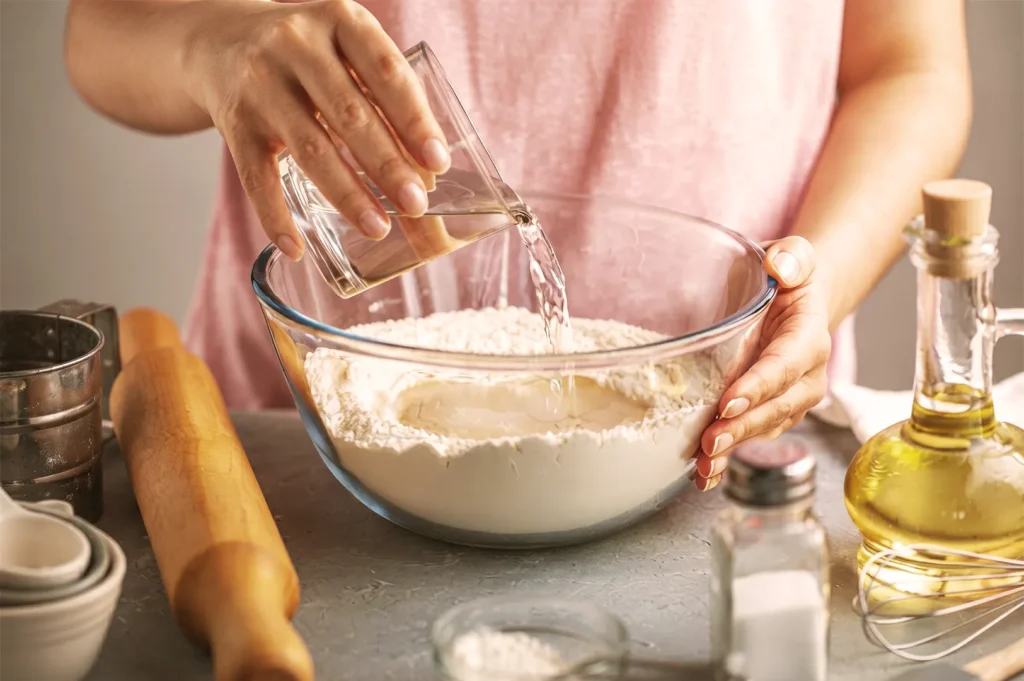 Hands pouring water from a glass measuring cup into a bowl of flour to prepare homemade dough.