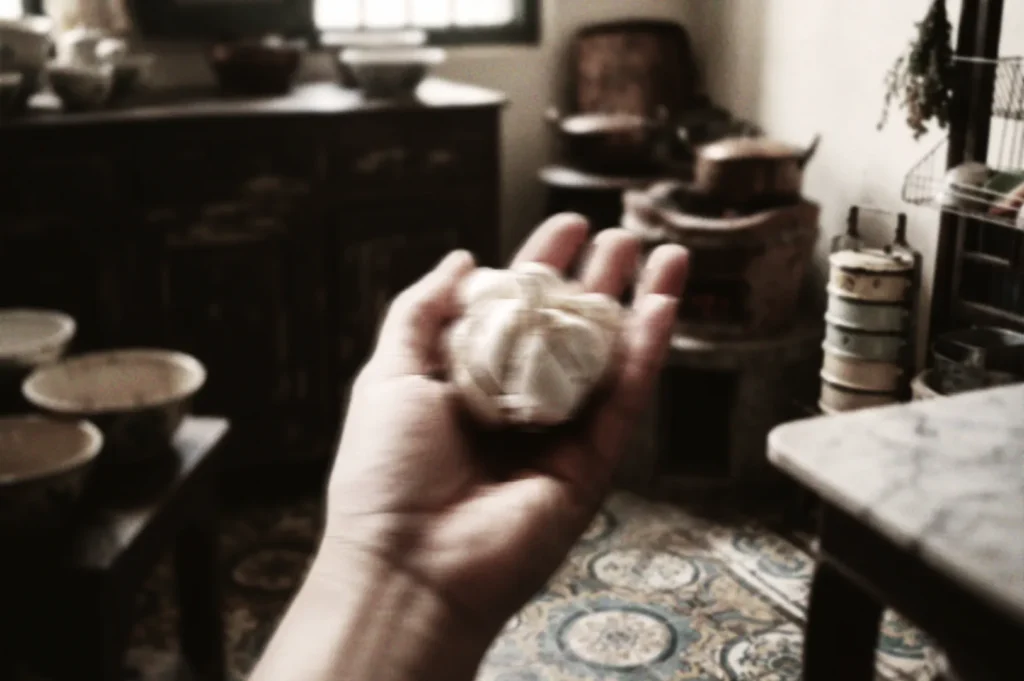 Close‑up of a hand holding garlic bulb in a rustic kitchen with patterned tiles and antique cookware, evoking traditional Asian cooking.