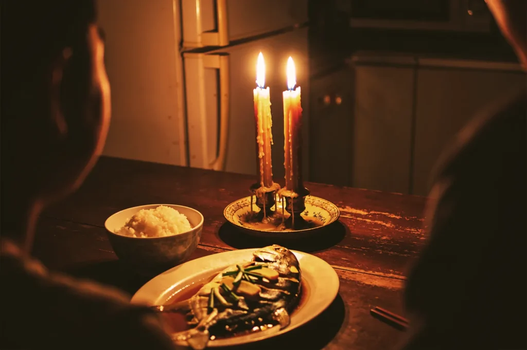 Two burning candles with wax drippings illuminating a plate of steamed whole fish and rice on a dark wooden table.