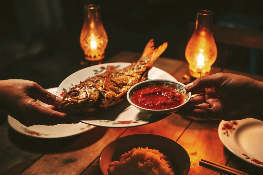 Hands passing a plate of crispy fried whole fish and a bowl of red chili sambal sauce across a wooden table lit by oil lamps.