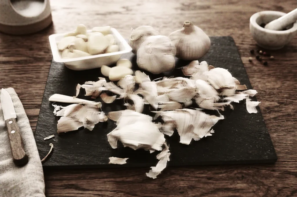 Fresh garlic cloves and peels on a black cutting board with knife and mortar, representing home cooking and natural ingredients.